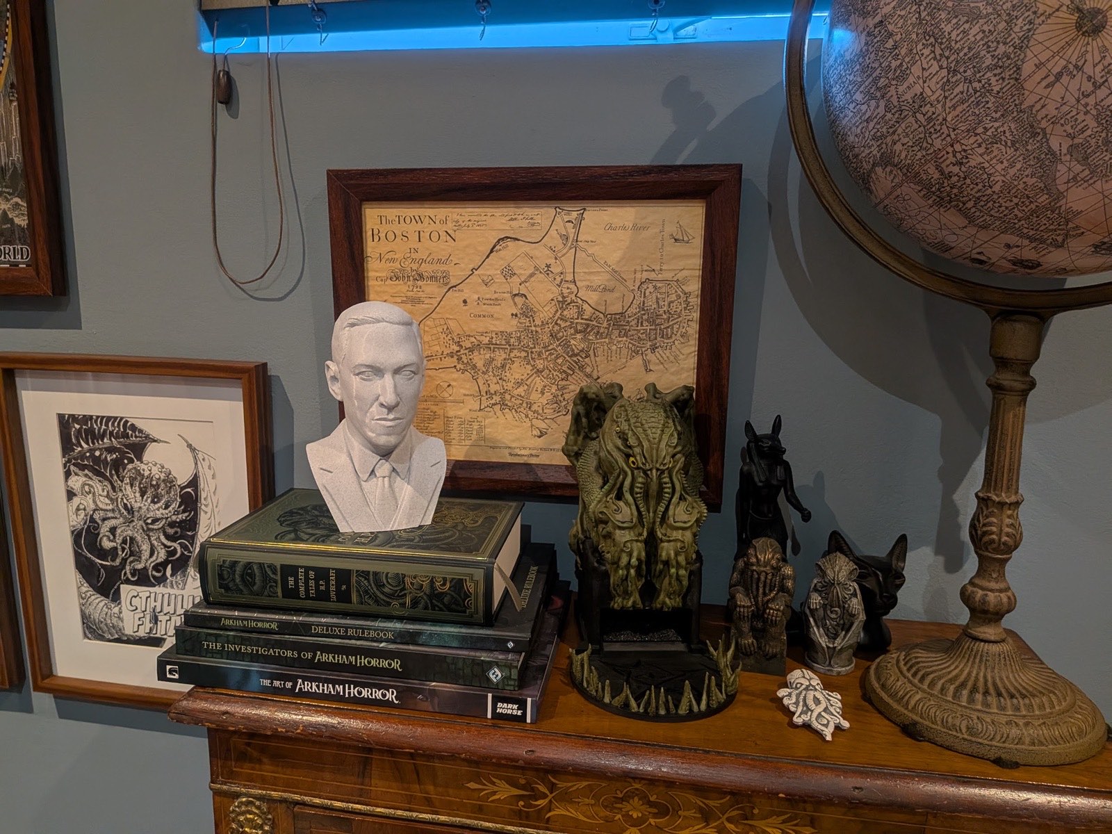 A bust of H. P. Lovecraft on a stack of Arkham Horror books, beside a Cthulhu idol and an antique globe